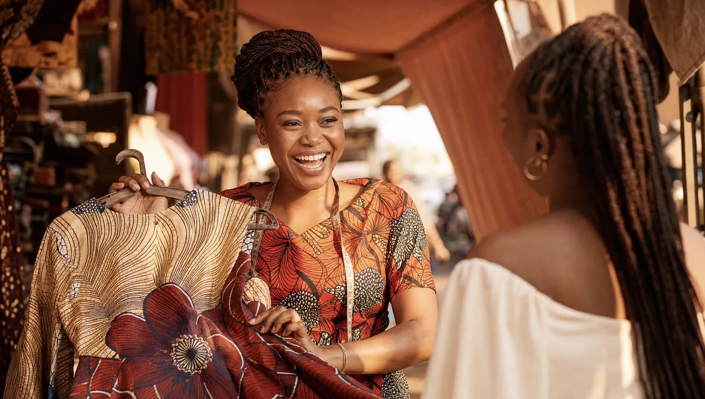 A Lagos fashion vendor laughing as she shows a dress to a customer at her sunlit ankara stall